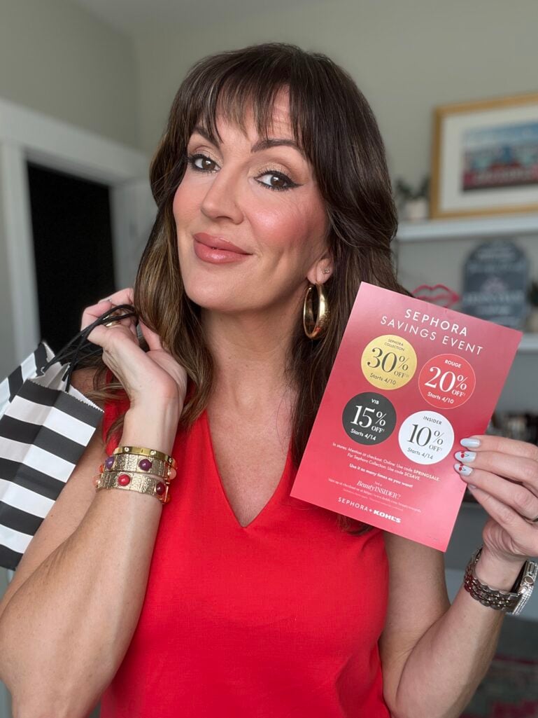 Woman with highlighted brown hair wearing red V-neck sleeveless top holding Sephora Savings Event flyer showing Rouge 20% off, VIB 15% off, and Insider 10% off discount tiers, with black and white striped Sephora shopping bag over wrist, gold jeweled cuff bracelet and silver watch bracelet, white nails with red cherry accent