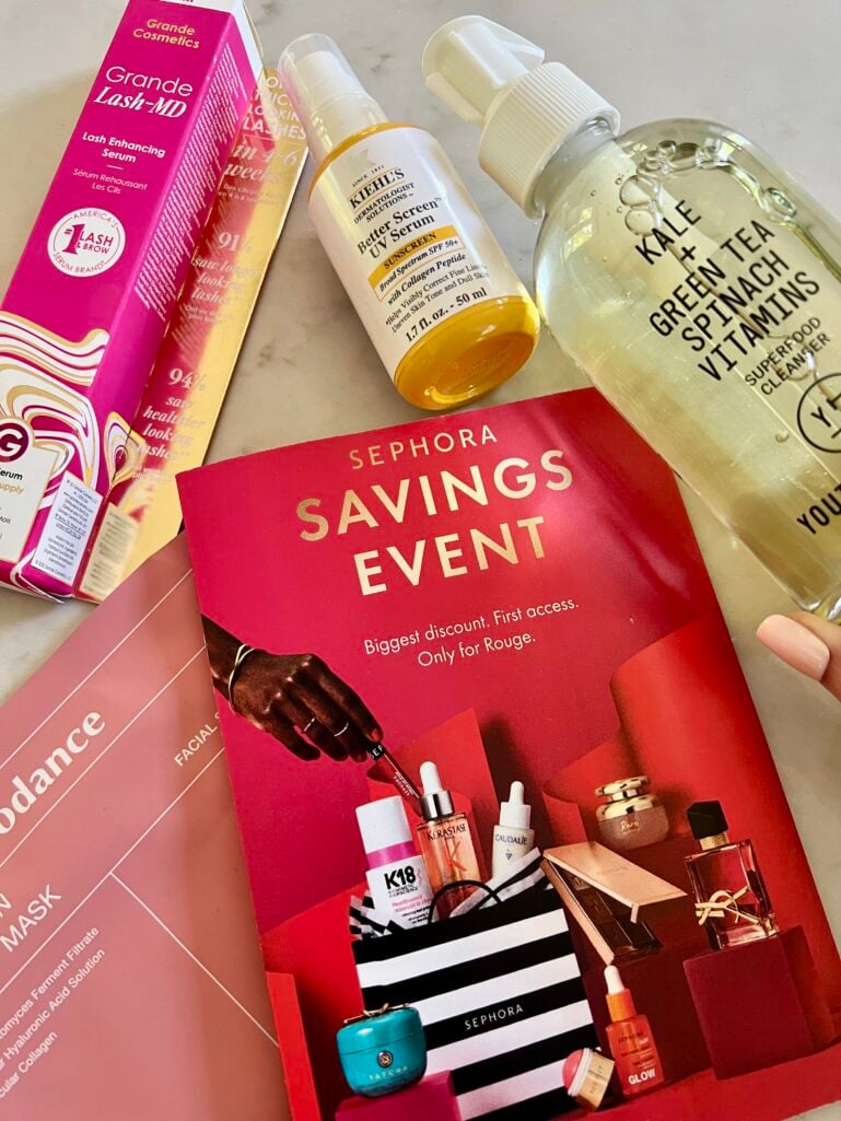 Flat lay product photo on white marble surface showing Grande Lash MD Lash Enhancing Serum pink box, Kiehl's Better Screen UV Serum with SPF 50 amber bottle, Youth to the People Superfood Cleanser clear green bottle, and red Sephora Savings Event brochure, Biodance mask packaging partially visible