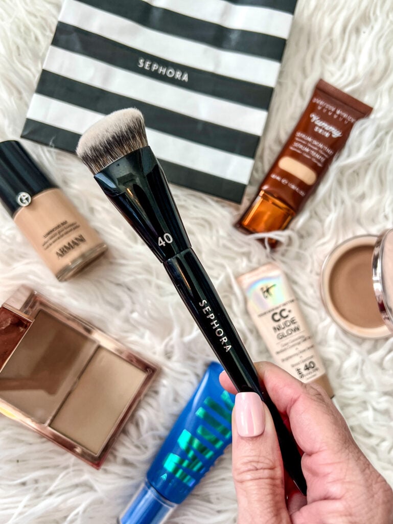 Hand holding Sephora Collection #40 Pro buffing foundation brush with black handle against white fur background, surrounded by beauty products including Armani Beauty Luminous Silk foundation, IT Cosmetics CC+ Nude Glow tube, NARS concealer pot, Charlotte Tilbury compact, and blue Urban Decay product, light pink manicure visible, black and white striped Sephora bag in background