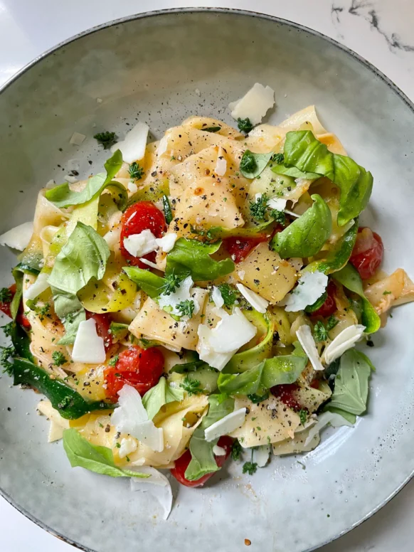 Overhead view of summer pasta with zucchini and tomatoes in a speckled gray bowl, topped with shaved Parmesan, fresh basil leaves, chopped parsley flowers, and fresh cracked black pepper on a marble surface