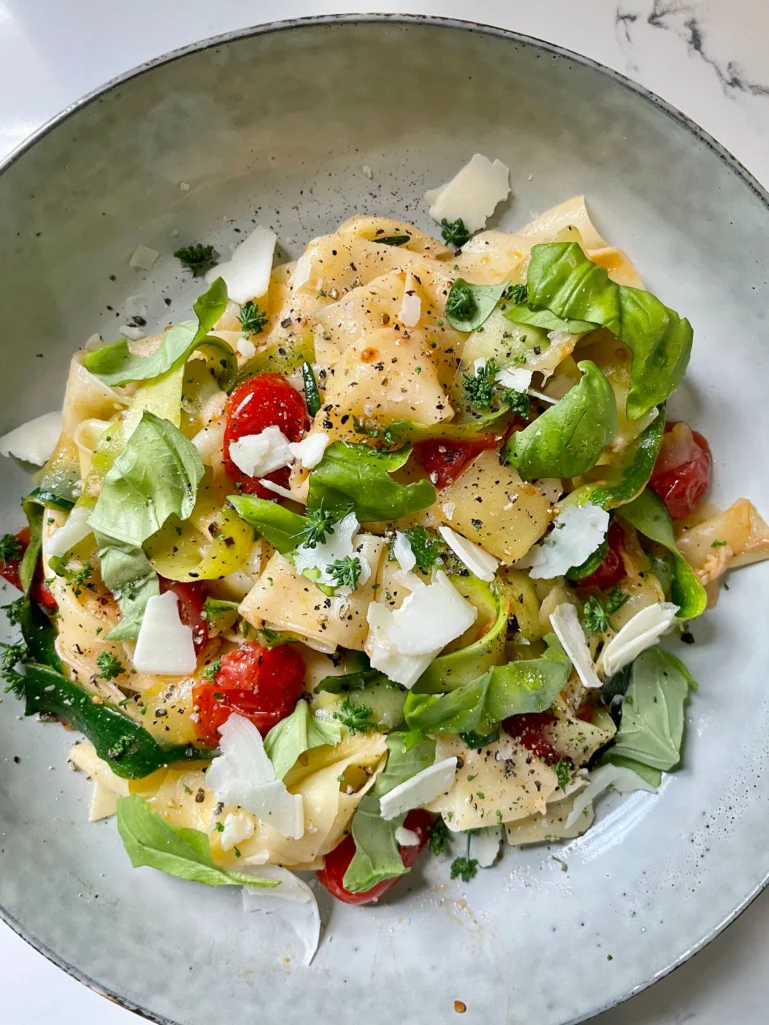 Overhead view of summer pasta with zucchini and tomatoes in a speckled gray bowl, topped with shaved Parmesan, fresh basil leaves, chopped parsley flowers, and fresh cracked black pepper on a marble surface