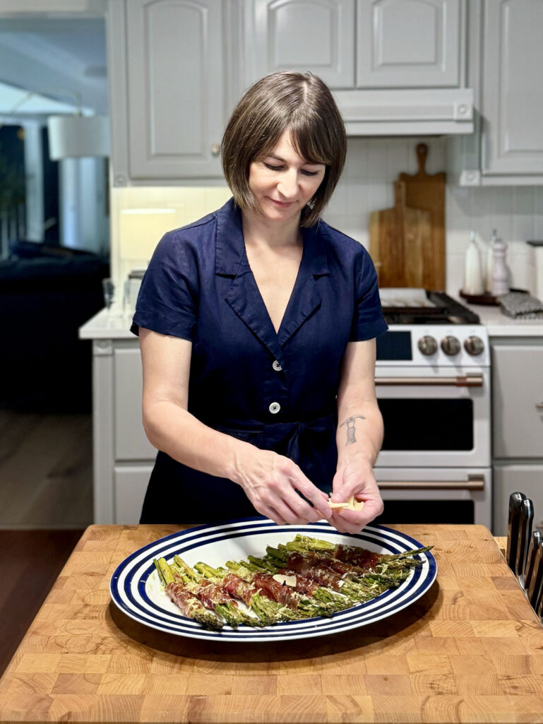 Kelly stands at a butcher block kitchen counter, smiling as she carefully adds shaved Parmesan to a platter of roasted asparagus and prosciutto bundles, wearing a navy linen shirtdress, with gray cabinets and a stainless range visible behind her.