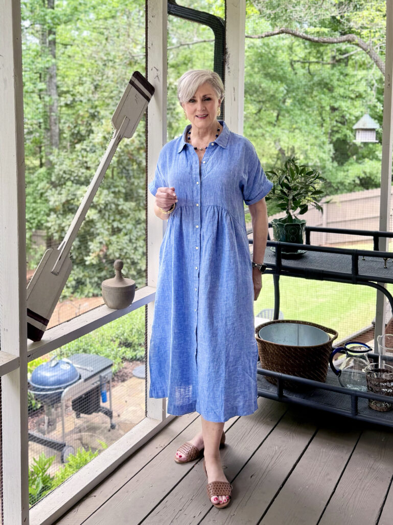 Beth stands on a screened porch in a periwinkle blue linen shirtdress with a smocked waist, woven leather slide sandals, a beaded necklace, and gold hoop earrings, one hand raised, with lush green trees visible through the porch screens behind her.