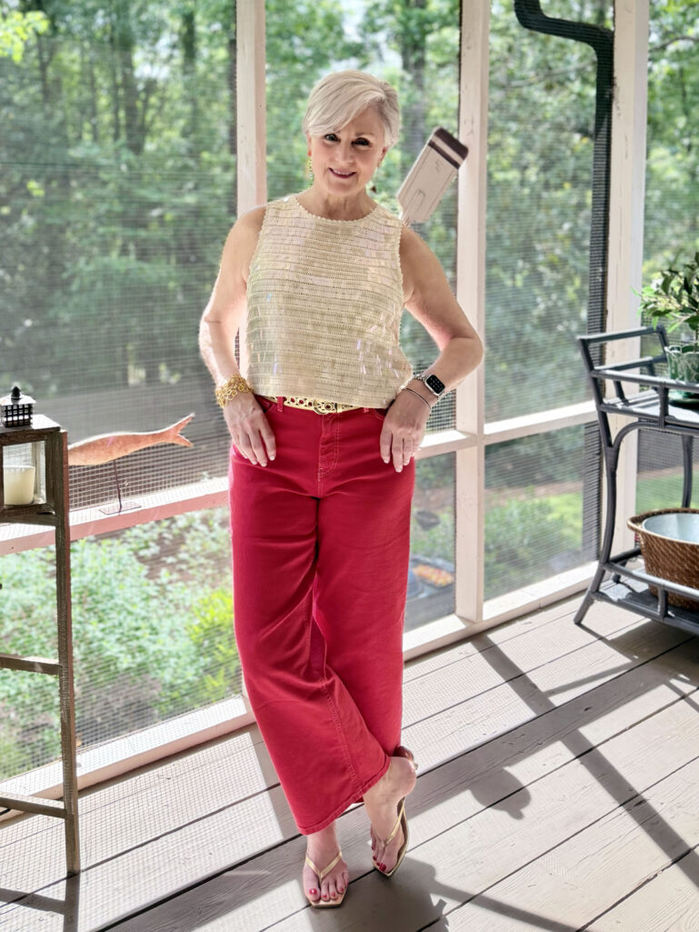 Beth stands on the screened porch in a cream iridescent sequin shell top, deep red wide-leg crop jeans with a gold braided leather belt, and gold thong kitten heel sandals, one hand on her hip, smiling at the camera with lush green trees visible through the porch screens behind her.