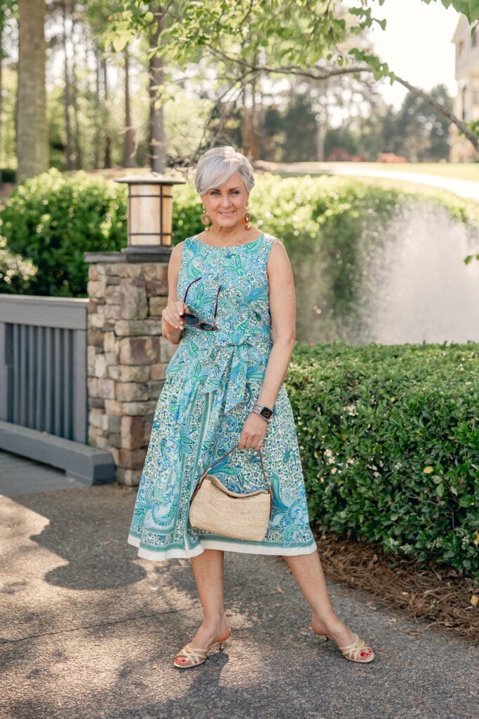 Woman over 50 with silver hair smiling outdoors in a blue and green paisley fit and flare midi dress, carrying a natural woven shoulder bag and holding tortoise sunglasses, wearing gold drop earrings and nude kitten mule sandals — a second styling view of the Mother's Day dress in regular sizing.