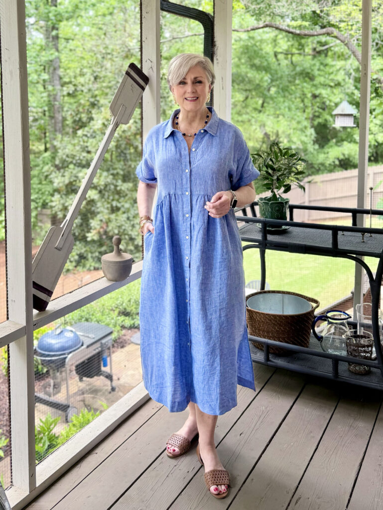 Full-length view of Beth on the screened porch in her periwinkle linen shirtdress and woven sandals, smiling with both hands clasped lightly in front, green trees and a bar cart visible through the porch screens in the background.