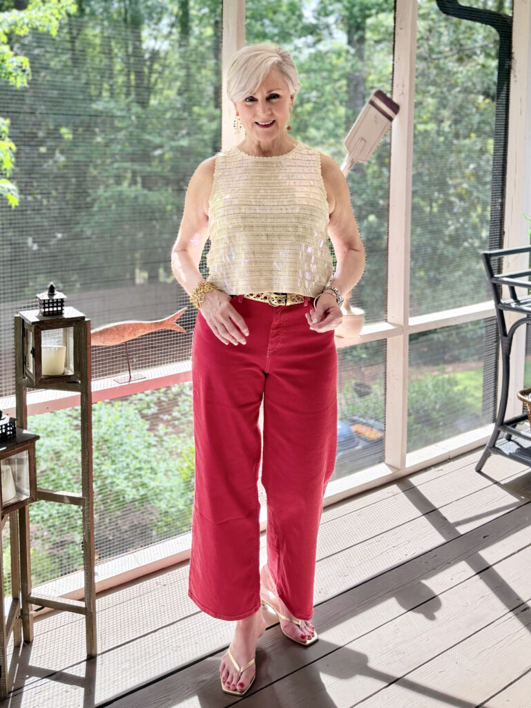 Full-length view of Beth on the screened porch in her sequin shell top, red wide-leg crop jeans, gold belt, and gold kitten heel sandals, both hands resting at her hips, with porch lanterns and greenery visible behind her.