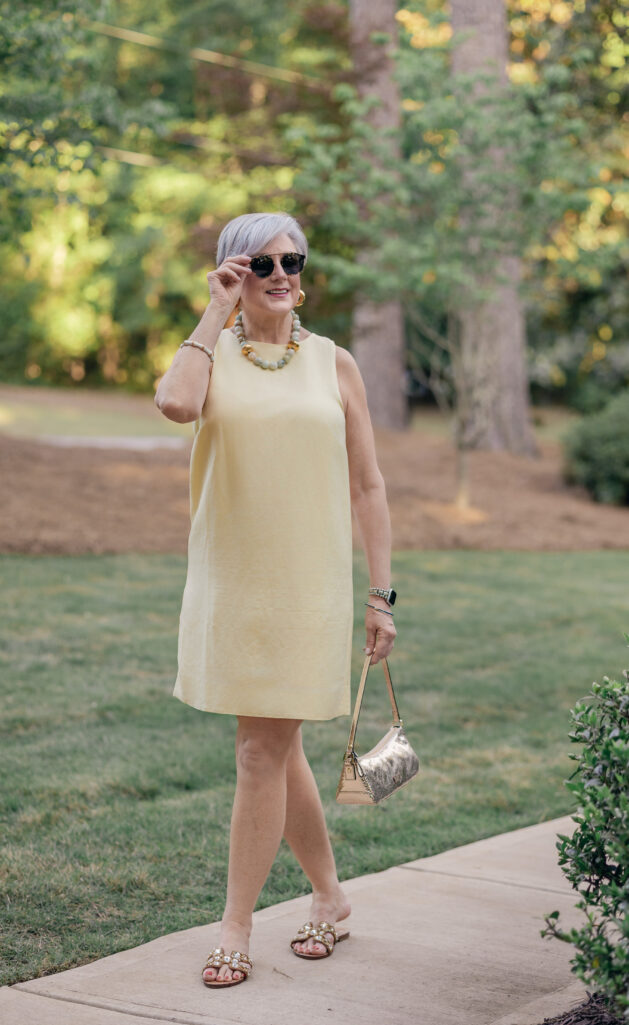 Summer 2026 colors after 50 — woman wearing butter yellow sleeveless shift dress with gold sandals and beaded necklace