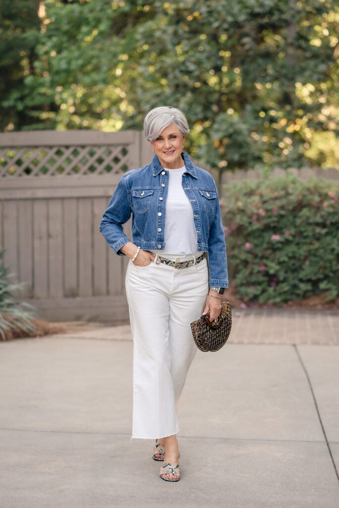 Woman over 50 with silver hair wearing a cropped denim jacket over a white tee with white wide-leg jeans, a snake-print belt, snake-print slide sandals, and a small dark woven handbag on a paved walkway — a classic summer outfit idea featuring white wide-leg jeans for women over 50.