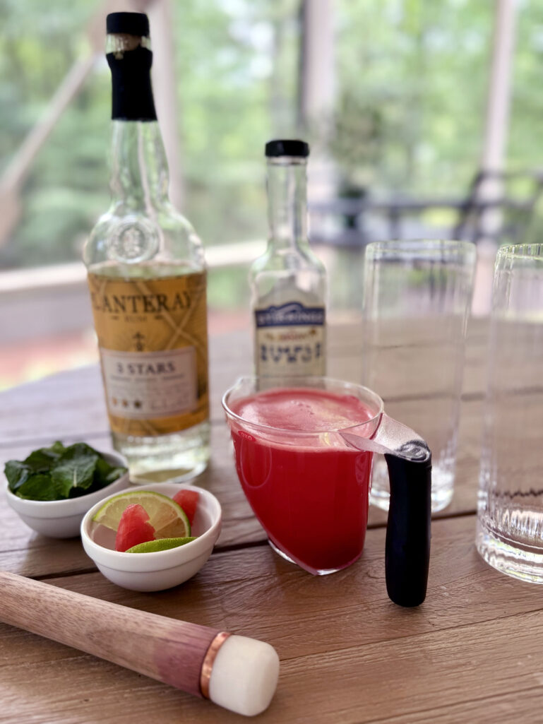 An outdoor table styled with a bottle of Plantation 3 Stars white rum, a bottle of simple syrup, a glass measuring cup filled with fresh blended watermelon juice, a small white bowl of fresh mint leaves, a white bowl of lime and watermelon garnishes, and a wooden muddler, green trees visible through the screened porch in the background.