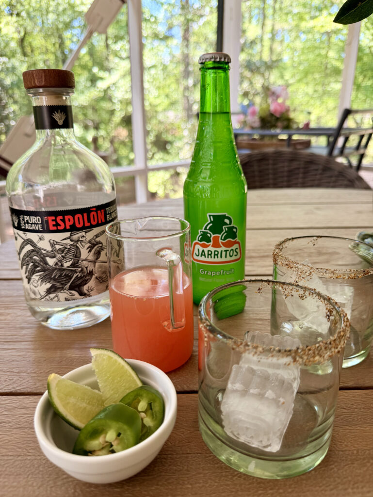 An outdoor table styled with a bottle of Espolón Blanco tequila, a bottle of Jarritos Grapefruit soda, a glass pitcher of pink grapefruit juice, two tajín-rimmed rocks glasses with large ice cubes, and a small white bowl of fresh jalapeño slices and lime wedges, lush green trees visible through the screened porch windows.