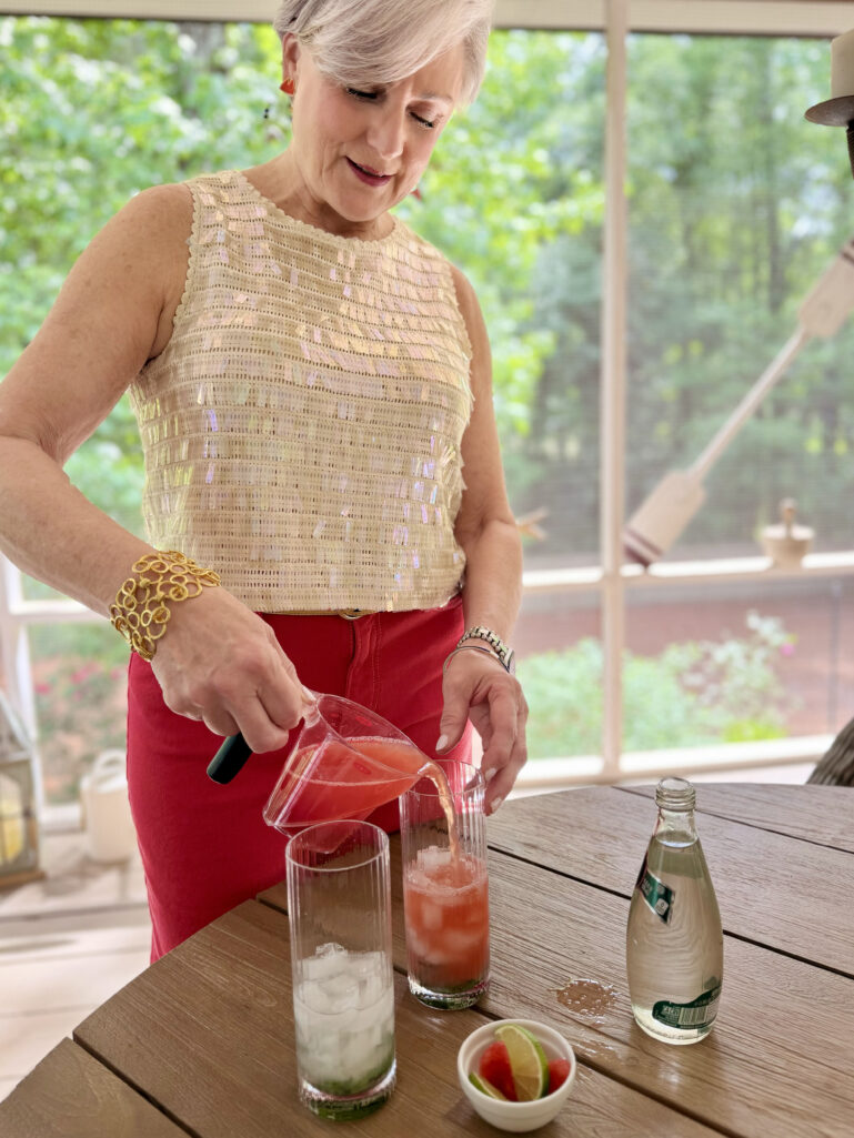 Beth leans over an outdoor table, pouring fresh blended watermelon juice from a glass measuring cup into two highball glasses filled with muddled mint and ice, wearing her cream sequin shell top and red wide-leg jeans, club soda and garnishes nearby on the table.