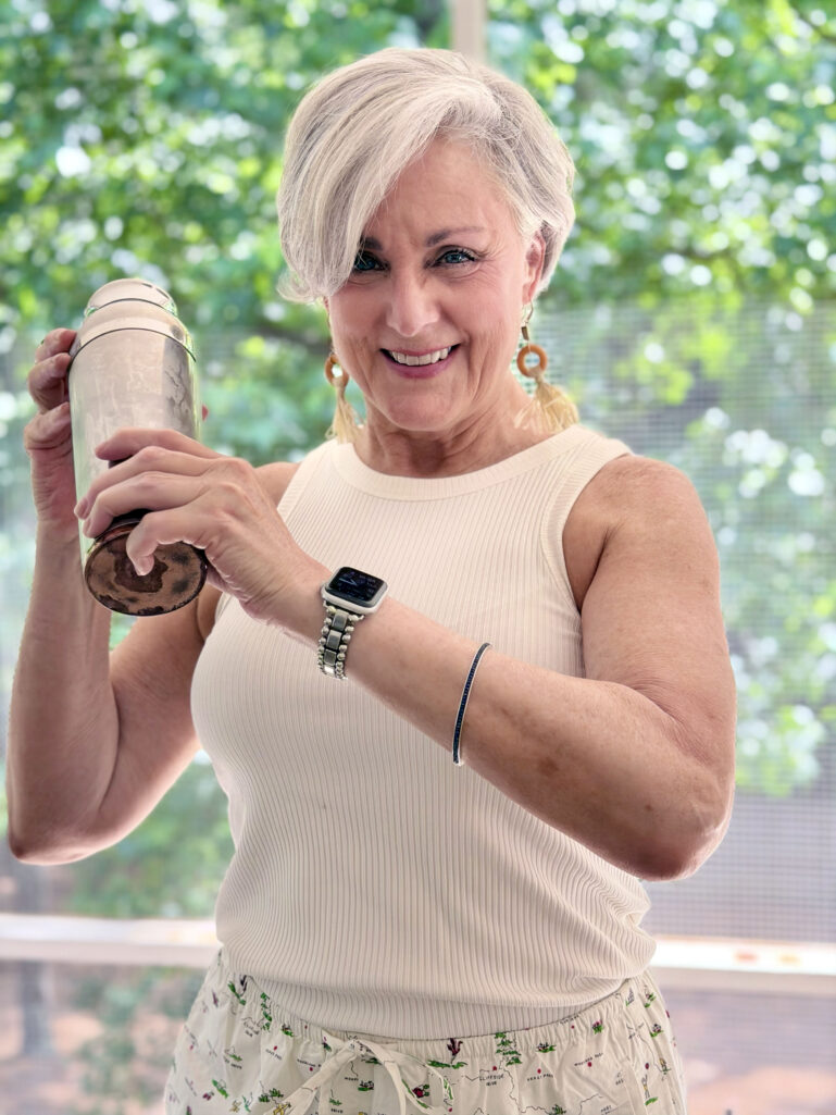 Beth in a cream ribbed tank top and tassel earrings smiling while shaking a silver cocktail shaker on the porch, with green foliage behind her.