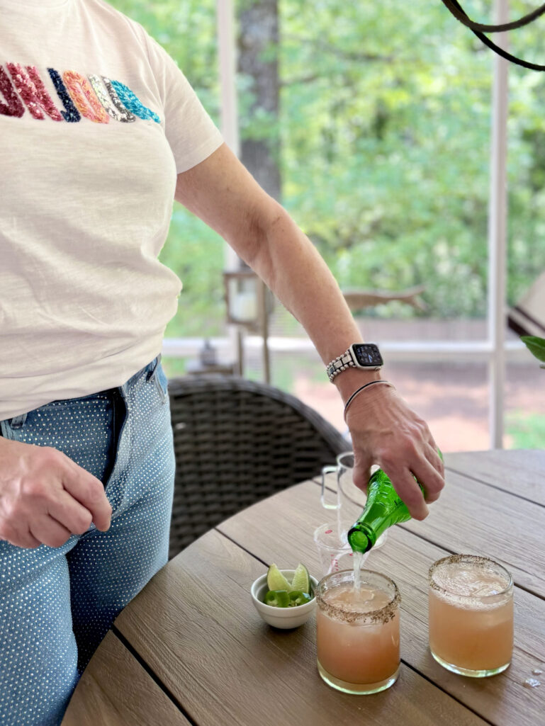 Beth leans over an outdoor table pouring Jarritos Grapefruit soda into two tajín-rimmed rocks glasses filled with spicy paloma, a small white bowl of jalapeños and lime wedges nearby, the screened porch and garden visible behind her.