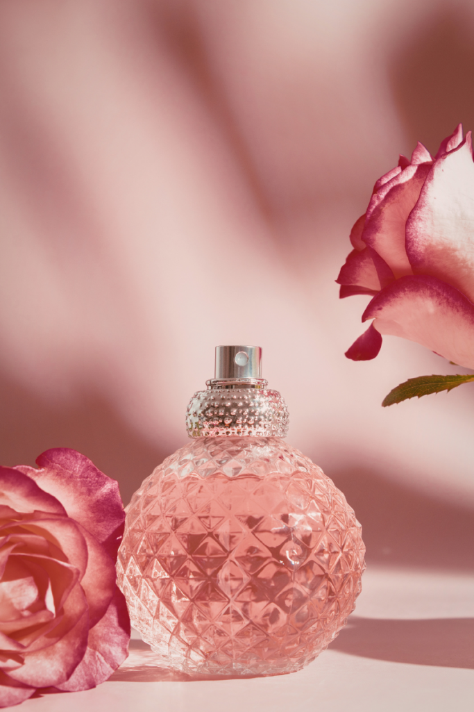 Romantic flat lay of a faceted pink glass perfume bottle surrounded by blooming roses against a soft blush background.