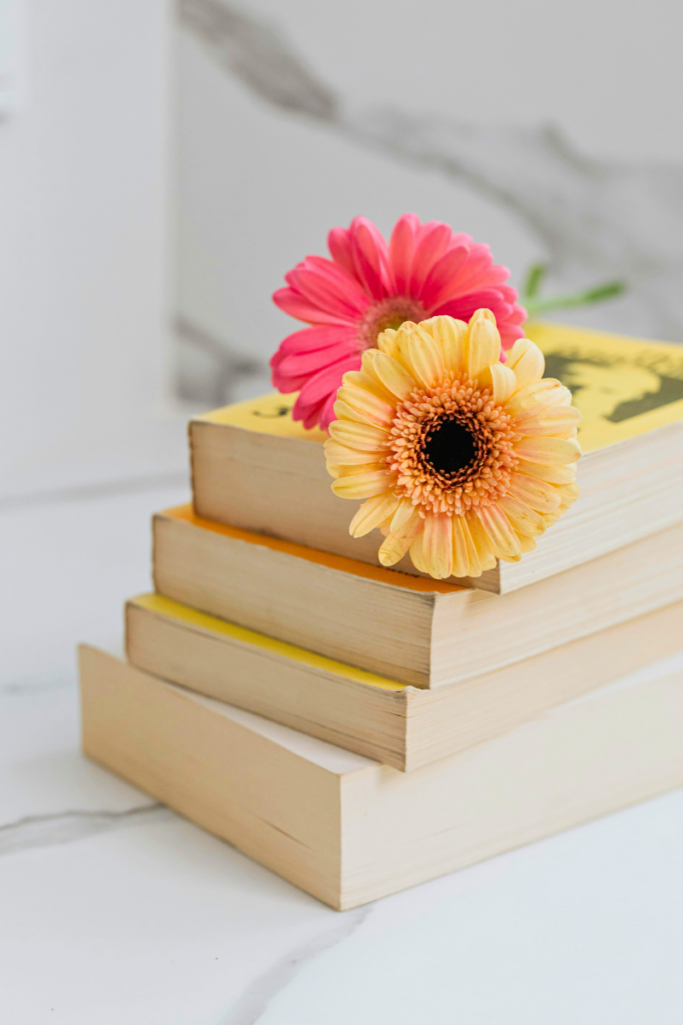 Stack of neutral-toned books topped with bright pink and yellow gerbera daisies on a white marble surface, creating a cheerful and cozy reading-inspired flat lay.