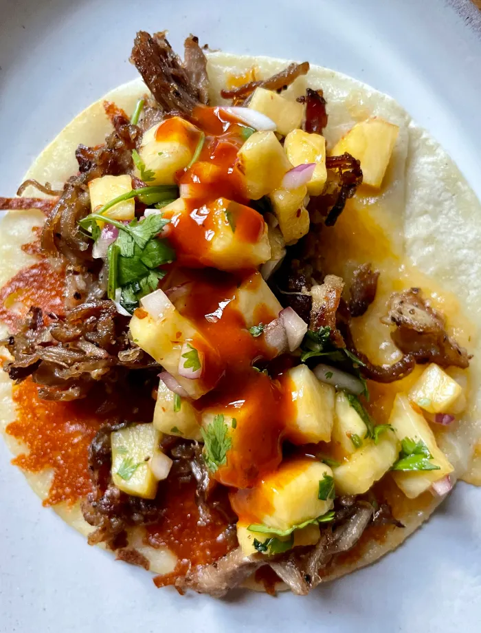 Close-up overhead of a single finished pork taco on a white plate topped with chipotle pineapple salsa and a drizzle of hot sauce over the crispy cheese-crusted corn tortilla