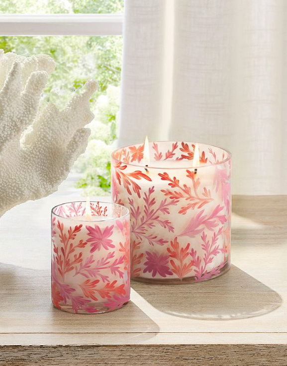 Sunlit vignette of pink floral glass candles glowing on a wooden surface beside a window, styled with soft linen curtains and coastal décor accents.