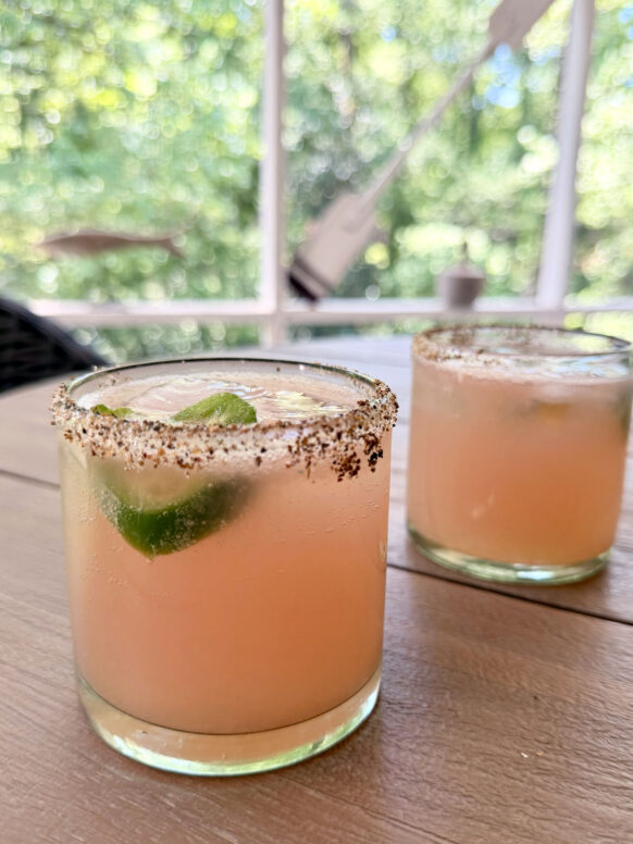 Two spicy paloma cocktails in tajín-rimmed rocks glasses sit on a wooden outdoor table, each garnished with a fresh jalapeño slice, with lush green bokeh trees visible through the screened porch behind them.