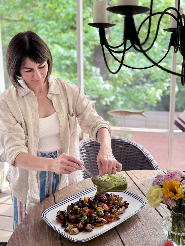 Kelly stands at the outdoor porch dining table, drizzling bright green chimichurri sauce from a small squeeze bottle over a platter of grilled chicken and vegetables, wearing her natural linen overshirt open over a white tank and colorful striped linen pants, a mason jar of summer flowers beside the platter.