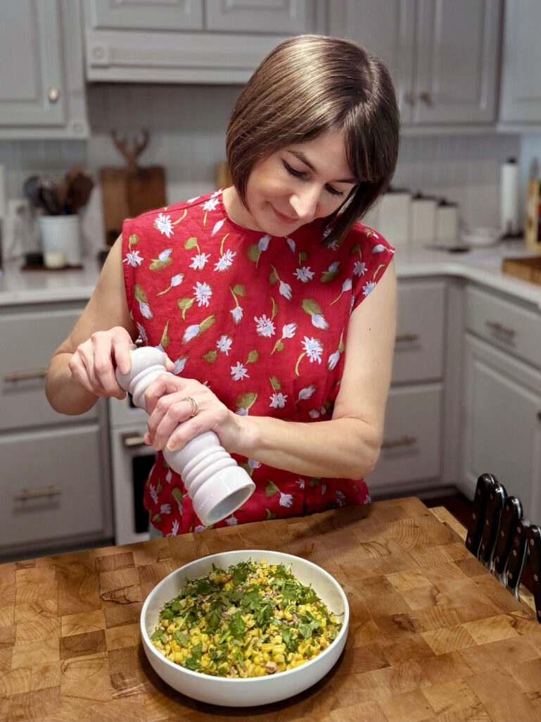 Kelly stands at a butcher block kitchen counter, smiling as she grinds fresh black pepper over a white bowl of street corn salad topped with fresh cilantro, wearing her vibrant red floral sleeveless top, with gray cabinets and kitchen tools visible behind her.