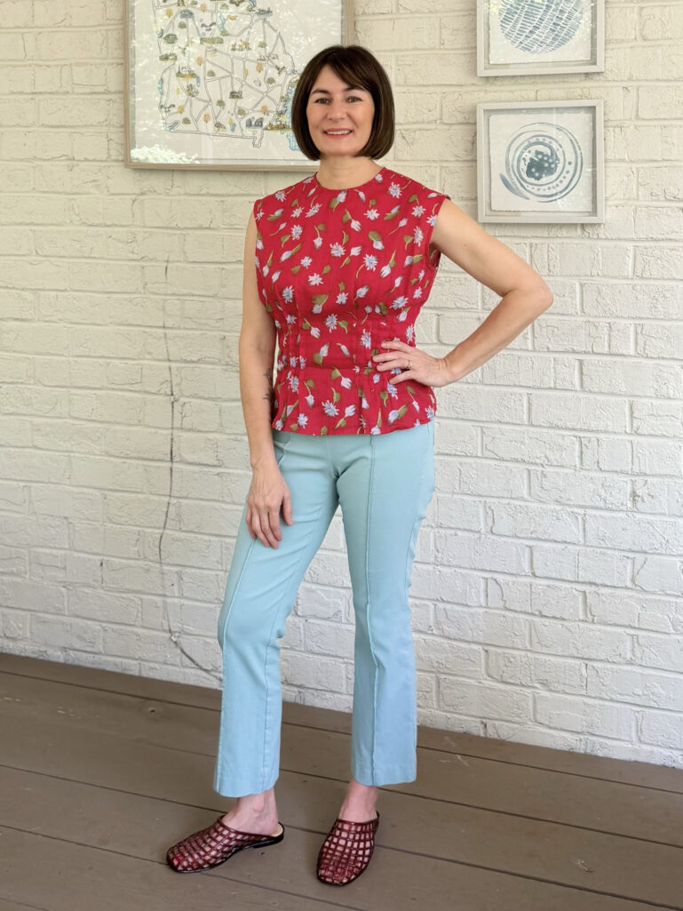 Kelly stands on the porch against a white brick wall, one hand on her hip, wearing a vibrant red floral sleeveless peplum top, aqua straight-leg Ivy pants with a front seam detail, and burgundy netted jelly mule sandals.