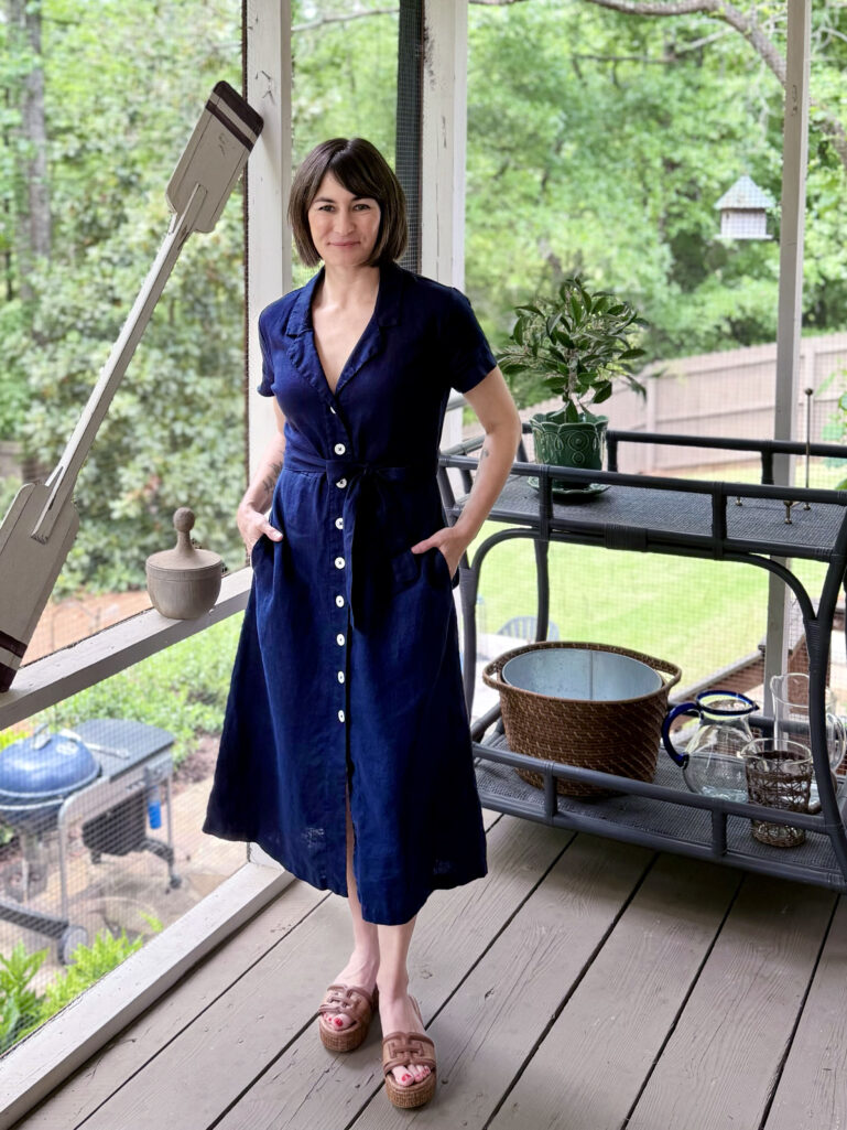 Full-length view of Kelly on the screened porch in her navy linen shirtdress belted at the waist, both hands in her pockets, one foot slightly crossed, smiling at the camera with lush green trees and porch details behind her.