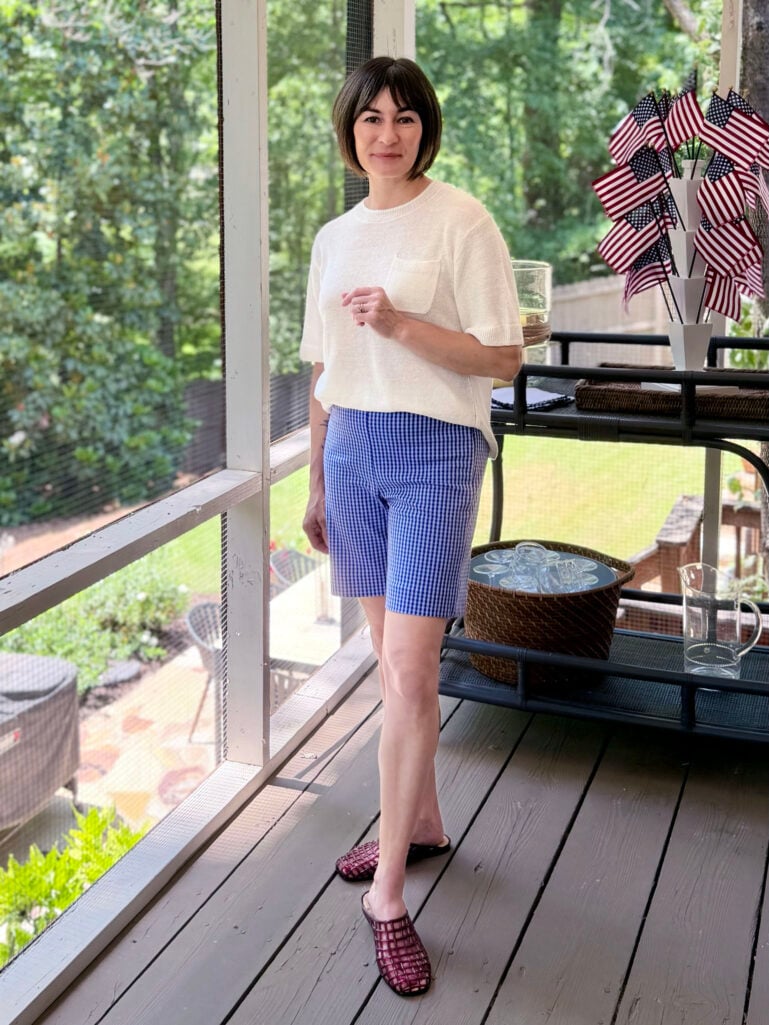 Kelly standing on the screened porch in a cream sweater tee and blue gingham Bermuda shorts with red jelly mules, one hand at her waist, American flags and a bar cart behind her.