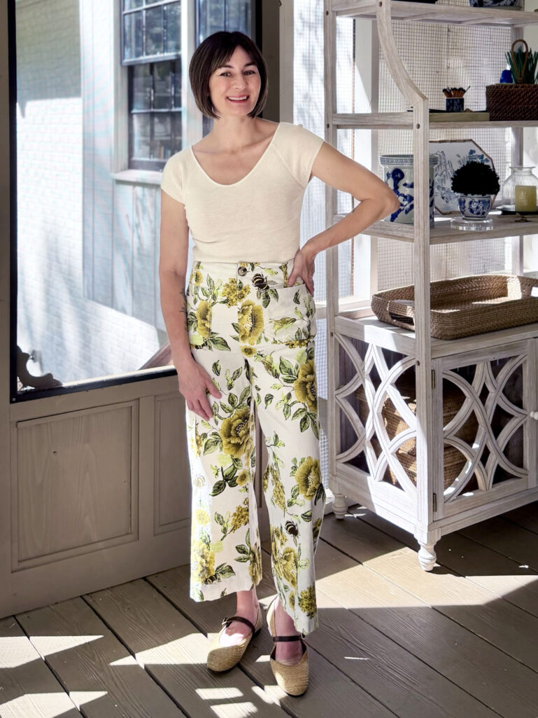 Full-length view of Kelly on the screened porch in a cream cashmere tee and floral wide-leg pants with raffia flats, smiling with windows and a baker's rack behind her.