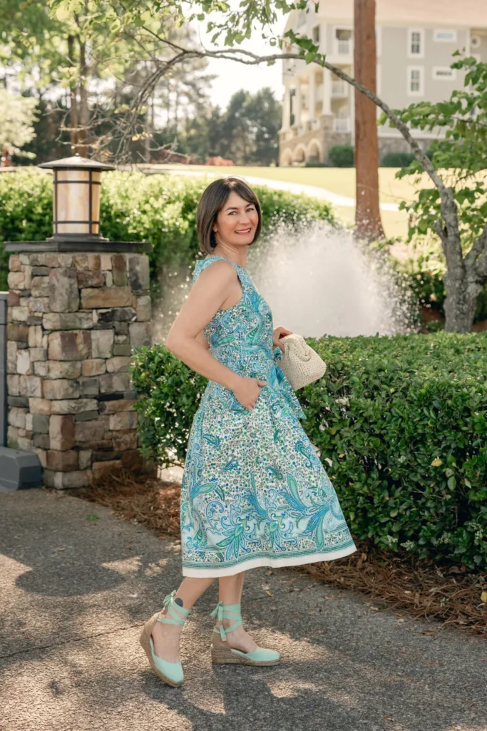Petite woman over 50 with brunette bob wearing a sleeveless blue and green paisley print cotton poplin fit-and-flare midi dress with mint green lace-up espadrille wedges and a small natural handknotted woven clutch — a vacation-ready summer cotton dress outfit idea for petite women over 50.