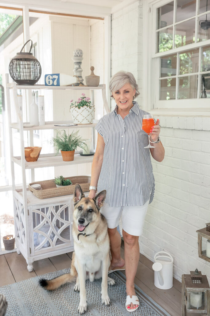 Beth stands on the screened porch in 2022, smiling and holding an orange cocktail, with Oscar — her large happy German Shepherd mix — sitting contentedly right beside her, the white porch shelving with decorative accessories and a window visible in the background behind them.