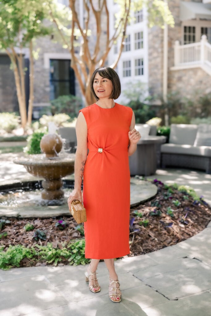 Kelly standing by a courtyard fountain in a fiery sunset-orange sleeveless midi sheath dress with a gathered twist waist detail, holding a wicker bee clutch and wearing gold strappy heeled sandals — petite sheath dress styled in a bold solid color