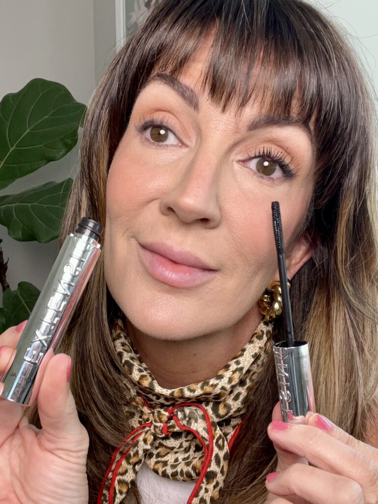 Woman holding L'Oreal Paris Extensionist lengthening and curling mascara wand, showing the dramatic difference between one eye with mascara on full curled lashes and one bare eye without, demonstrating the lash lifting effect, wearing leopard neckerchief and gold flower earrings