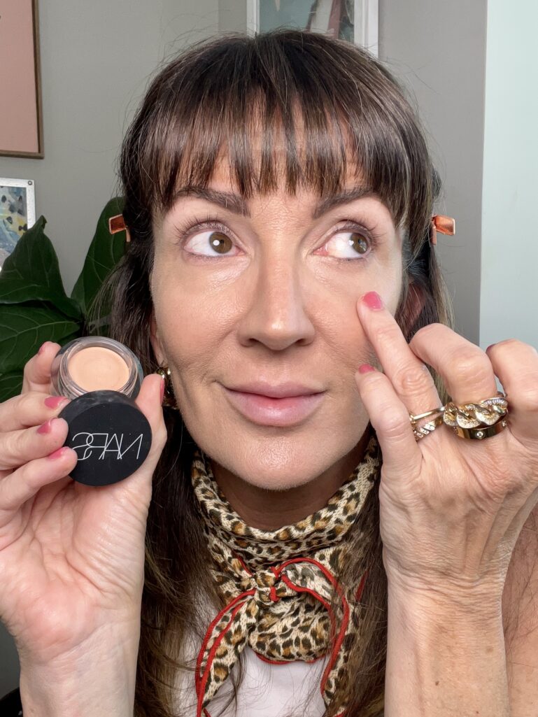 Woman applying Nars Soft Matte Complete Concealer in Creme Brulee under eye with finger, holding open round concealer jar showing nude shade, wearing leopard print neckerchief, gold chain rings, and rose gold hair clips, abstract artwork in background