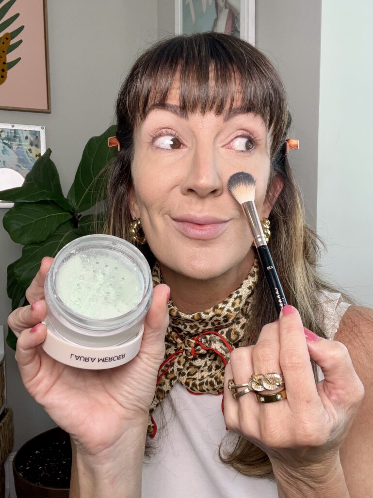 Woman applying Laura Mercier mint green color correcting loose powder with fluffy makeup brush to side of nose, holding open powder jar showing pale green powder, wearing leopard neckerchief and stacked gold rings, fiddle leaf fig plant visible in background