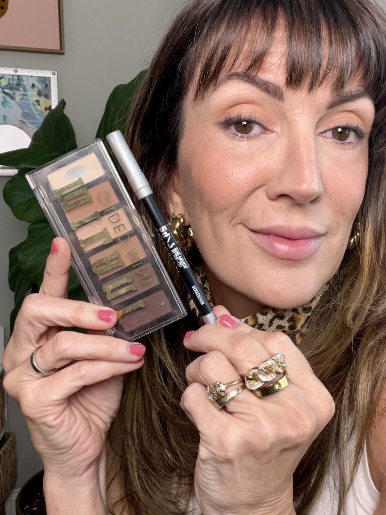 Woman holding Milani Gilded Mini eyeshadow palette in Whiskey Business showing neutral matte browns and Urban Decay 24/7 Glide-On Eye Pencil in Perversion black liner, wearing finished soft brown eye makeup, leopard neckerchief, gold chunky hoop earrings, and stacked gold chain rings