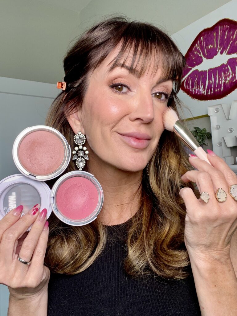 Woman applying cream blush with Rare Beauty Soft Pinch brush to high point of cheek, holding Makeup by Mario Soft Pop Plumping cream blushes in Barely Blushing neutral shade and Perfect Pink, wearing crystal earrings and stacked druzy rings