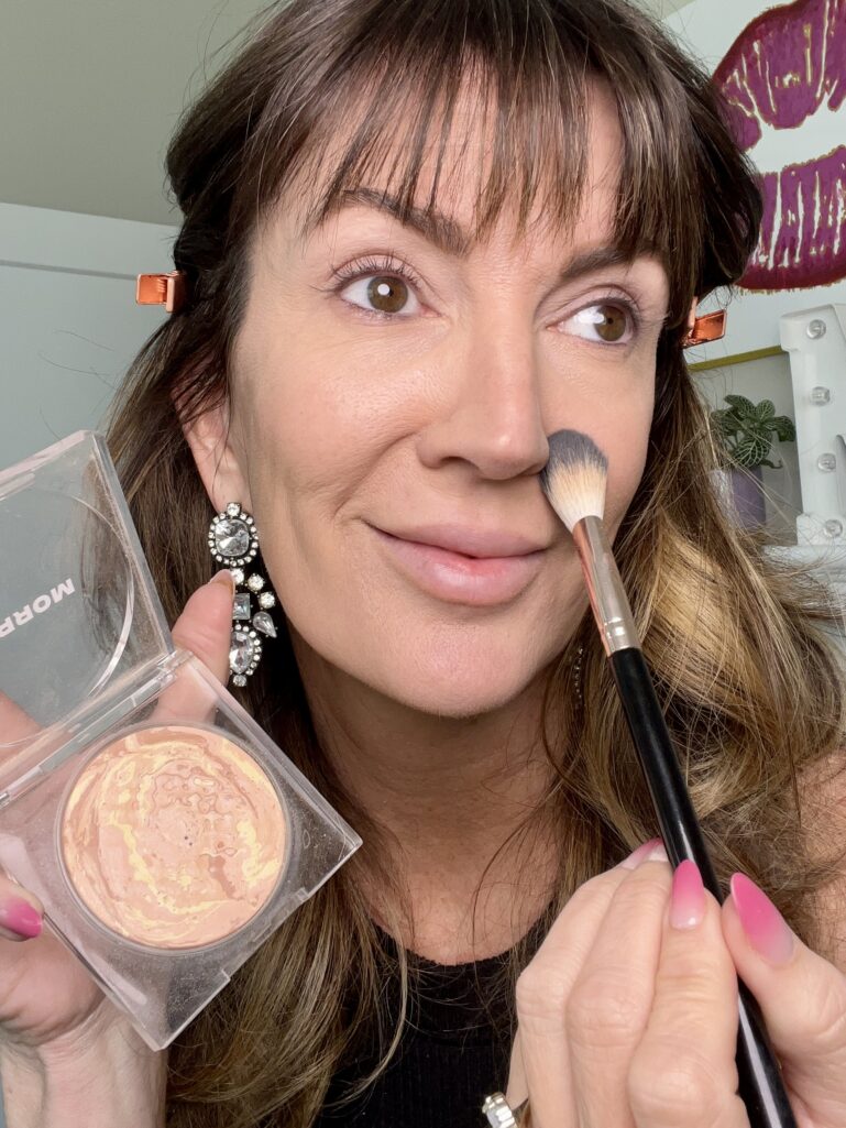 Woman applying Morphe Cloudlight Soft Glow veil powder in shade Sunrise with fluffy makeup brush to side of nose, holding clear powder compact, wearing crystal drop earrings and rose gold hair clips with finished foundation base