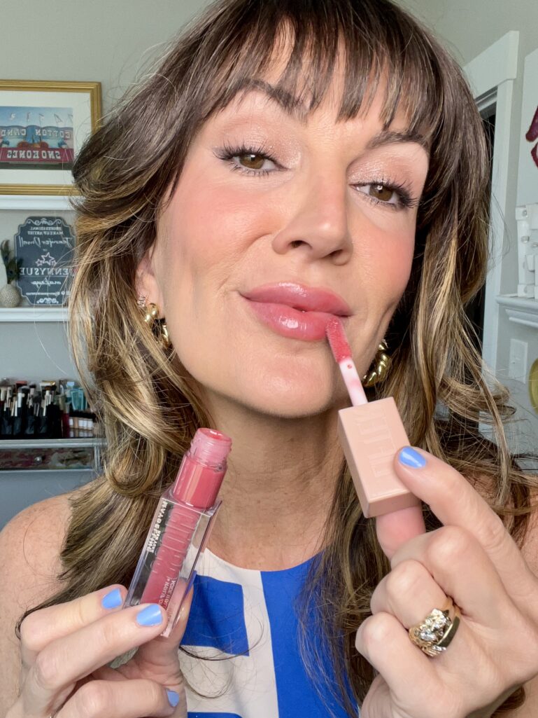 Close-up of a woman applying Maybelline Lifter Gloss in shade Heat to her lips with the doe-foot applicator, holding the pink-tinted tube, wearing soft glam makeup and periwinkle blue nails