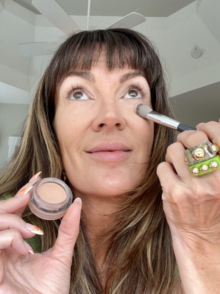 Woman applying Nars Soft Matte Complete Concealer in shade Creme Brulee under her eye with a small fluffy brush, holding the open pot of concealer, wearing chunky gold and green rings