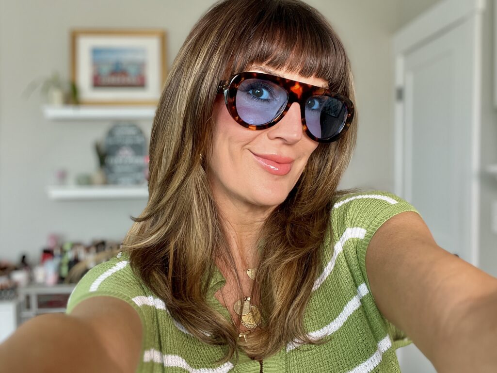 Woman taking a selfie wearing oversized tortoiseshell aviator sunglasses with blue-tinted lenses, glossy nude lips, and a green and white striped knit top