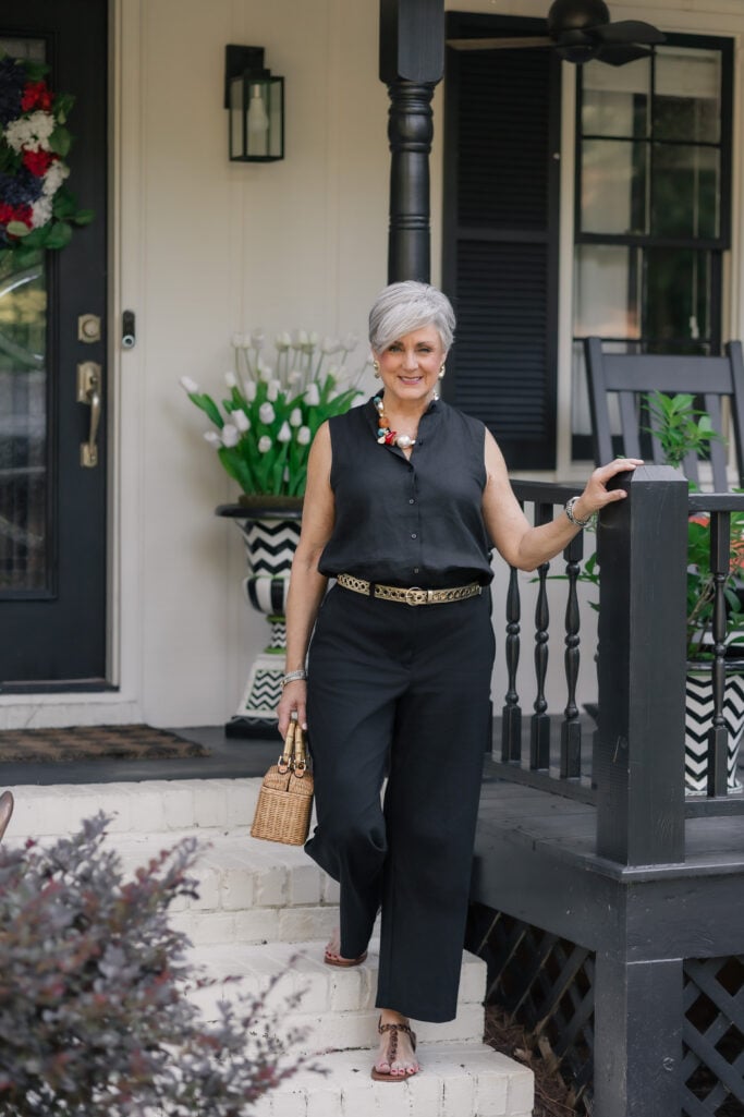 Woman over 50 on a black-and-white porch in a black sleeveless linen button-up top and wide-leg trousers from Eileen Fisher, with a gold openwork belt, pearl-and-stone statement necklace, beaded sandals, and a bamboo-handled wicker basket bag.