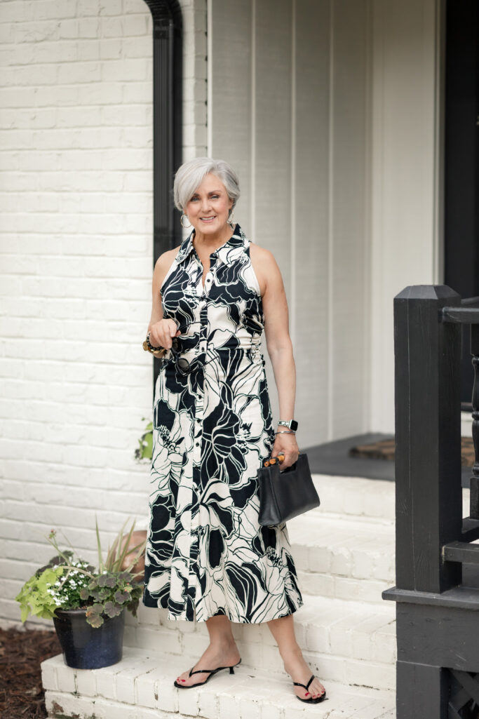 Beth wearing a black and white botanical floral print sleeveless shirtdress with black sandals and a black bag]