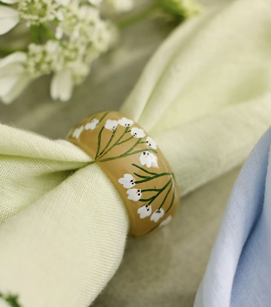 Hand-painted glass napkin rings in soft blue, pink, and amber hues, decorated with delicate floral motifs and displayed around pastel linen napkins on a seasonal tablescape.