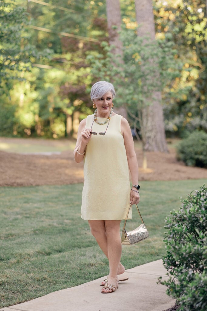 Beth wearing a pale yellow sleeveless linen shift dress with a beaded necklace and jeweled flat sandals
