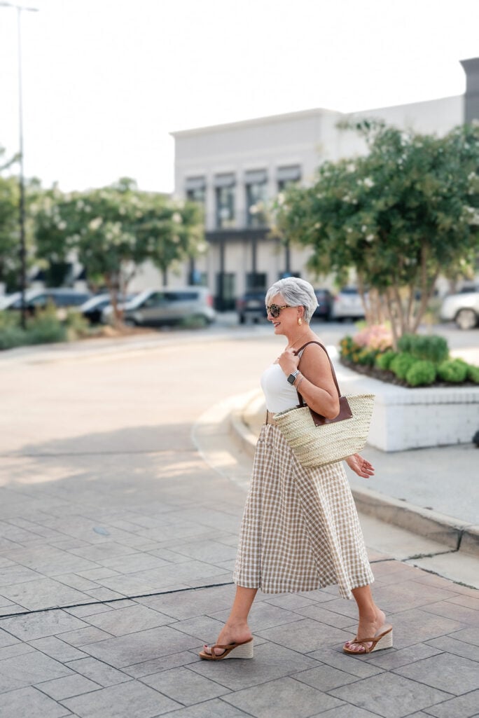 Woman with silver pixie cut walking in a tan gingham midi dress and straw tote — fall transition style