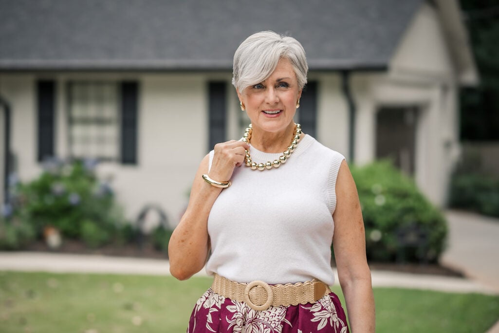 Beth of Style at a Certain Age facing forward and smiling, wearing a chunky gold ball bead necklace, small gold stud earrings, and a gold dome cuff bangle, styled with a white sleeveless sweater tank, burgundy floral midi skirt, and a woven raffia belt with a round wooden buckle
