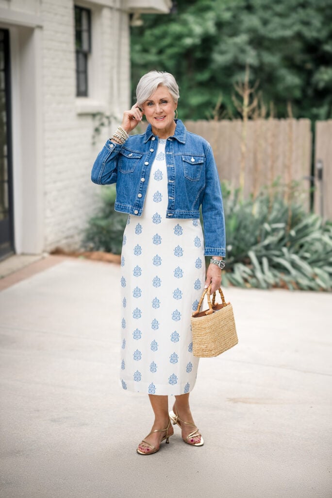 Beth of Style at a Certain Age in a denim jacket over a white and blue block print midi dress, right hand raised to ear, wearing a pearl and beaded bracelet stack on her right wrist and Apple Watch with link band on her left