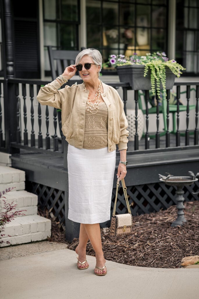 Beth of Style at a Certain Age in a tan linen bomber jacket over a gold textured cami and white linen pencil skirt, wearing a warm layered bracelet stack with pearls, gold bangles, and beaded pieces on her right wrist, holding sunglasses, with a Coach crossbody bag