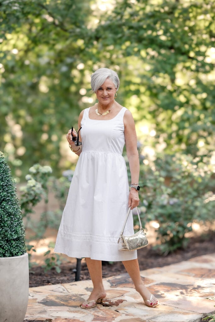 Beth wearing a sleeveless white cotton summer dress for women over 50 with a square neckline and gold accessories